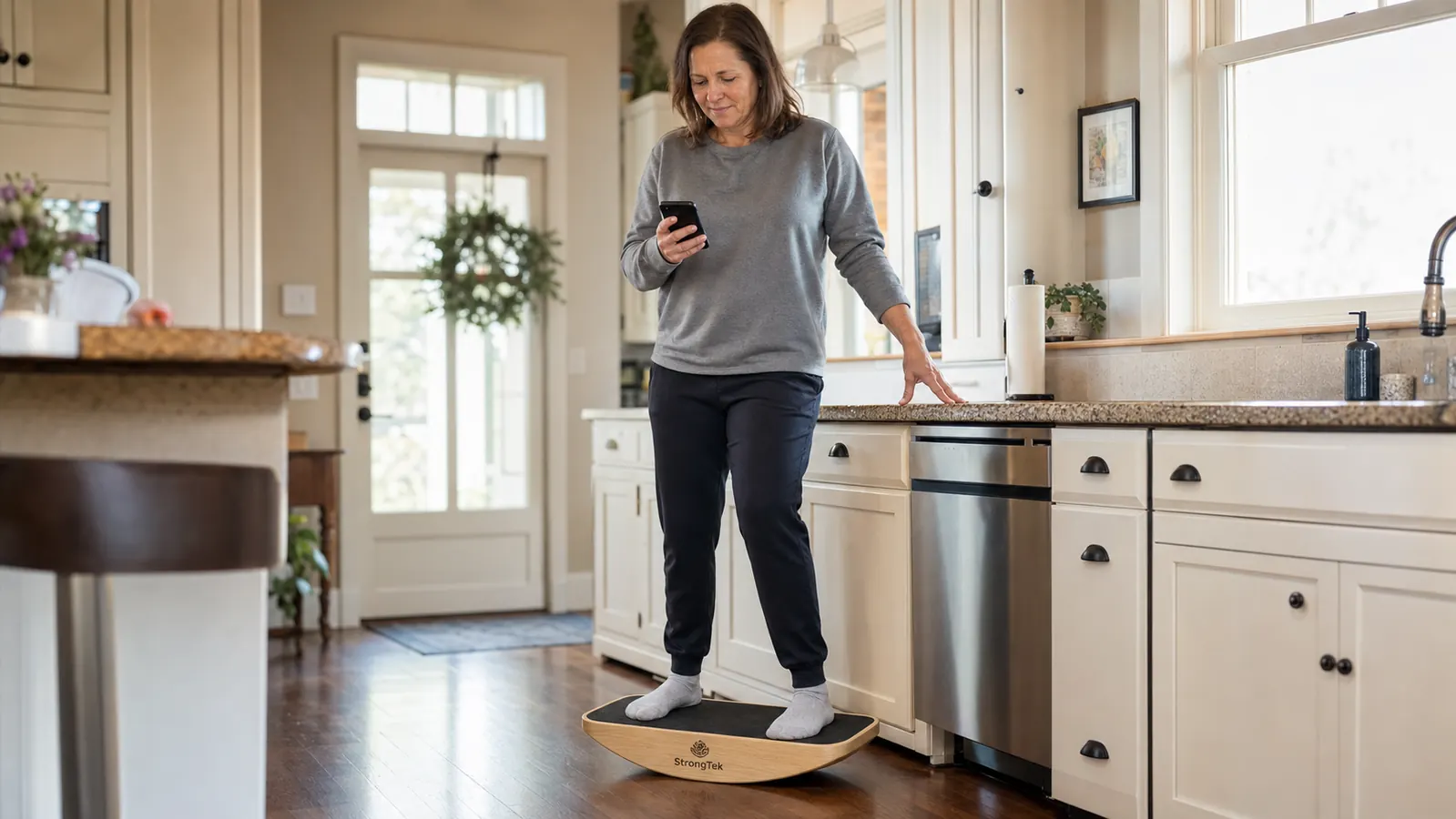 Woman in her fifties standing on a wooden balance board in a kitchen while holding a phone