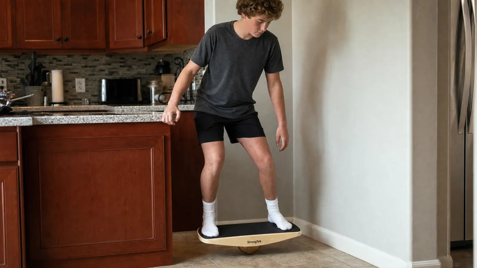 Teenage boy standing near a kitchen counter with one hand resting on the edge, balancing on the wobble board in a safe starting position