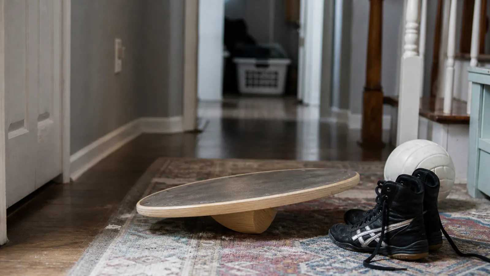 Wooden balance board sitting on a hallway rug next to a pair of wrestling shoes and a volleyball, morning light through a window