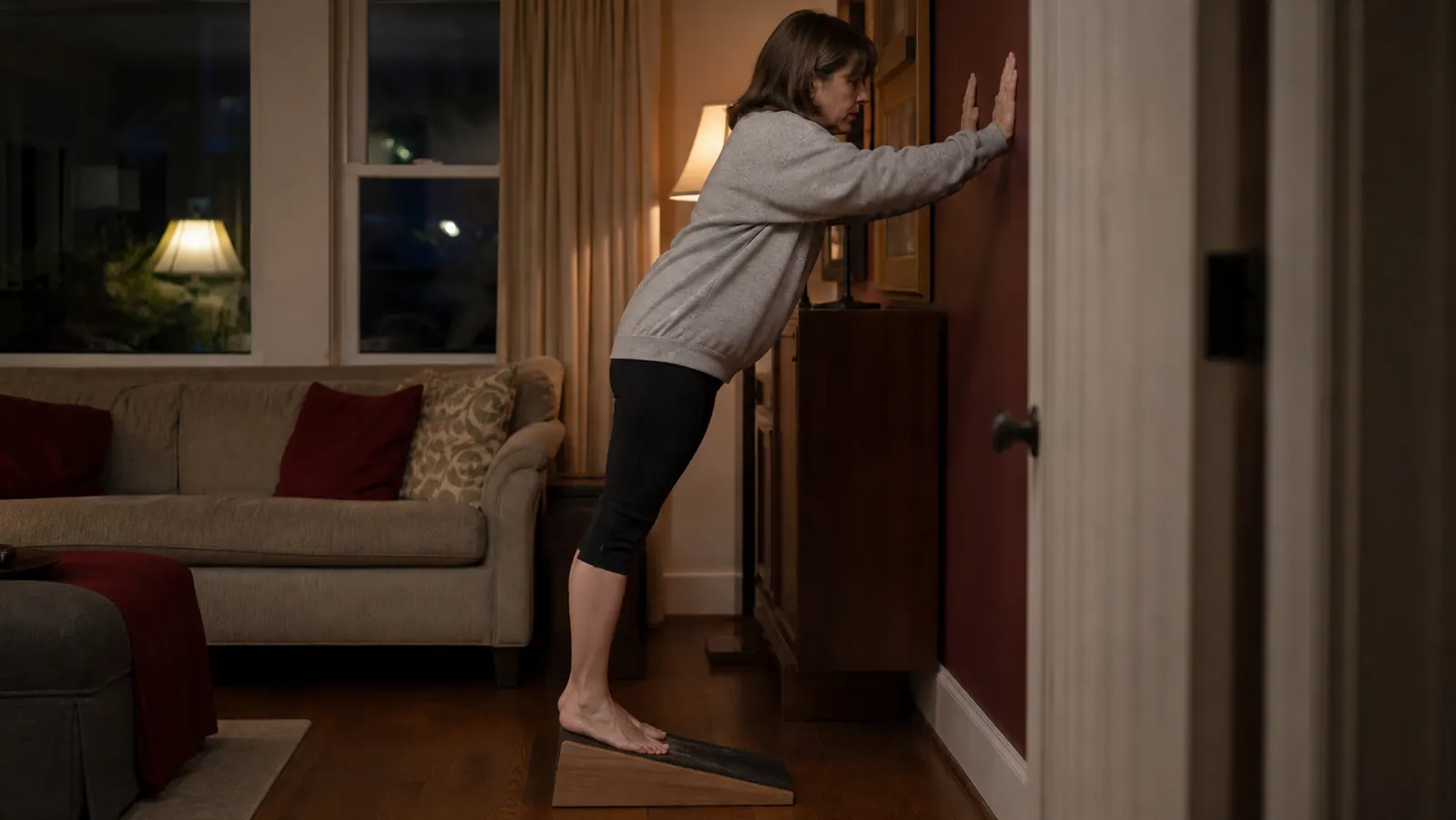 A woman in her fifties standing on a fixed angle slant board in a living room, leaning forward to stretch her calf in the evening