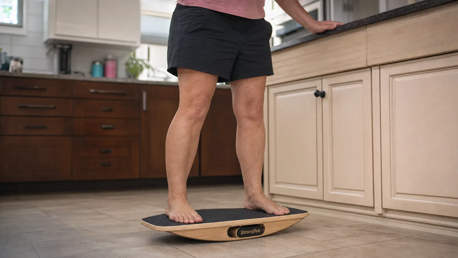 Close up of bare feet planted on a wooden balance board with a hand resting on a kitchen counter for support