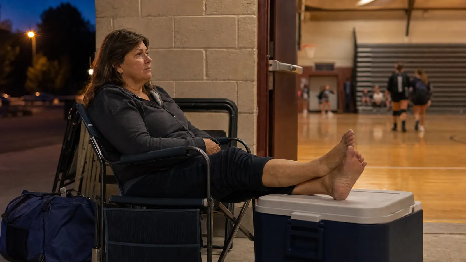 A woman sitting on a folding chair in a rec center parking lot with her shoes off, feet propped on a cooler, at the end of a tournament day