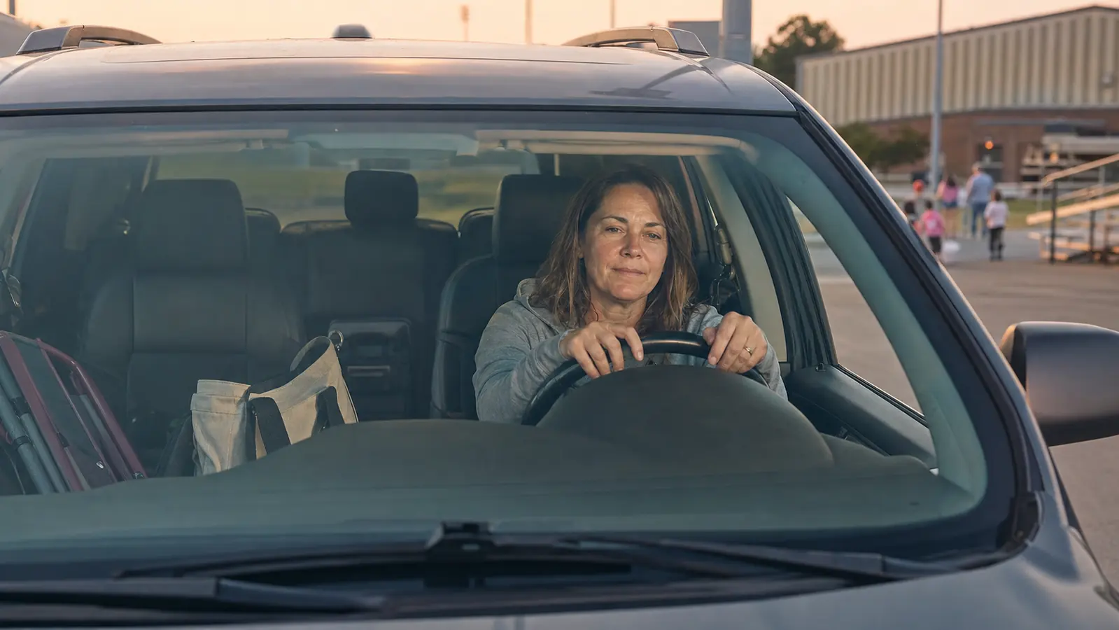 A woman in her fifties sitting in the driver's seat of an SUV at dusk, hands resting on the steering wheel, gym bag on the passenger seat