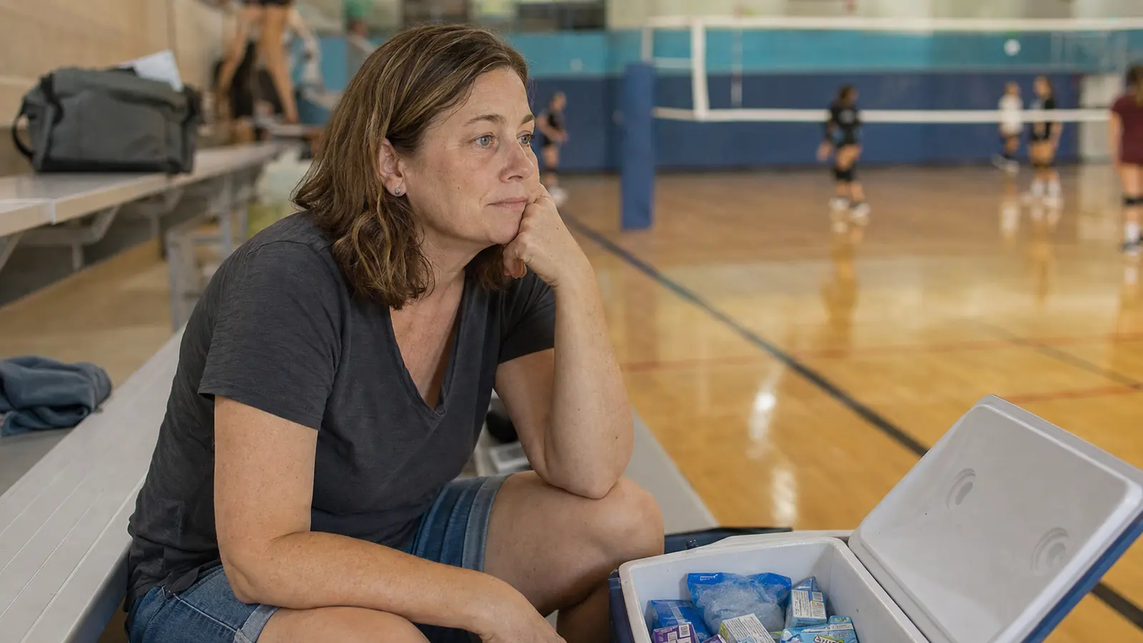 Woman in her fifties sitting on gym bleachers next to a loaded drink cooler, resting during a break in a volleyball tournament