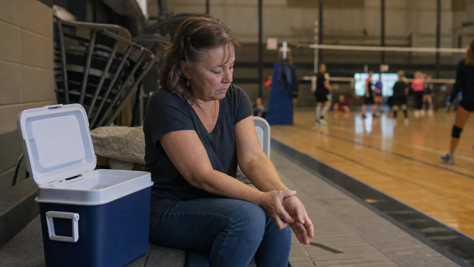 Woman in her fifties resting on gym bleachers with a loaded ice chest beside her, rubbing her wrist during a break in a volleyball tournament