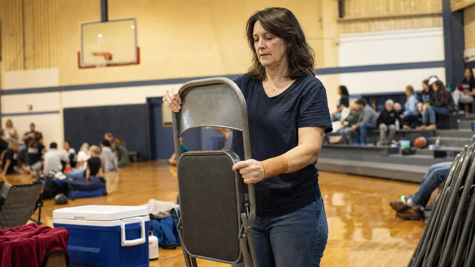 A woman folding a gym chair with a compression sleeve visible on her wrist, a loaded cooler beside her at a rec center gym